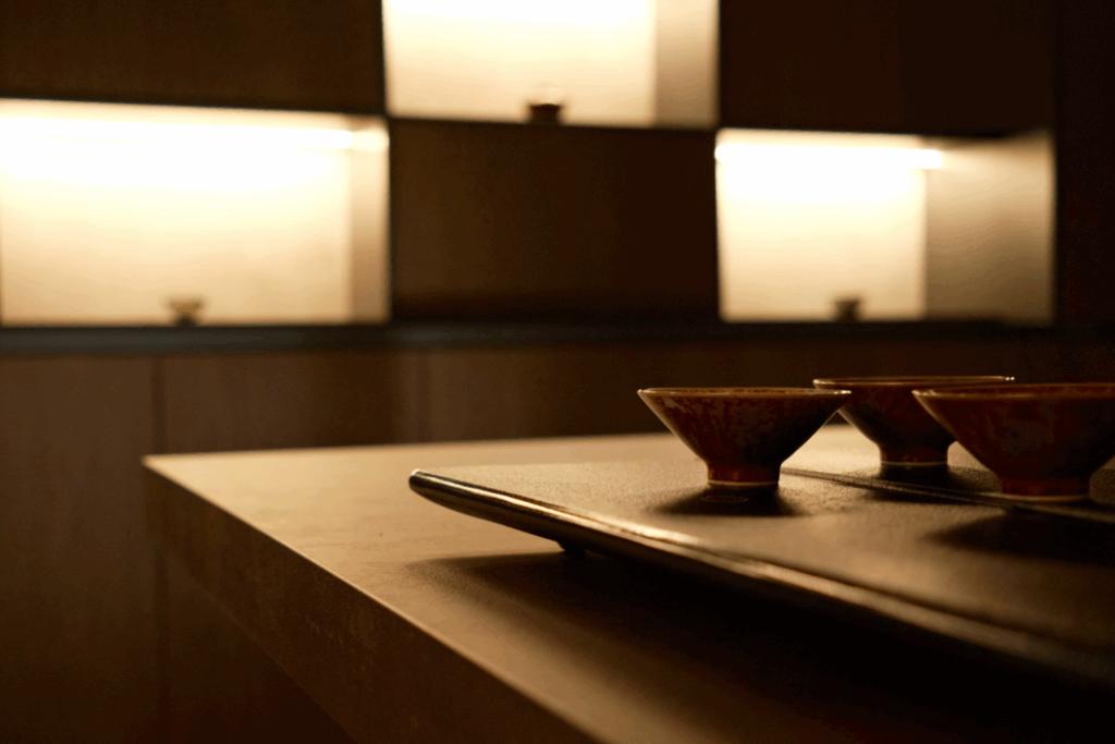 Three small ceramic tea bowls sit on a dark tray atop a wooden counter. The background features minimalist, softly illuminated recessed shelving that creates a warm and tranquil atmosphere.