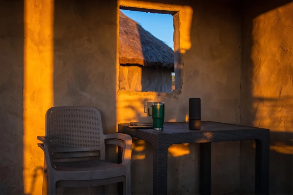 A green insulated tumbler and black speaker on a table inside a rustic hut during golden hour with a thatched roof visible through the window.