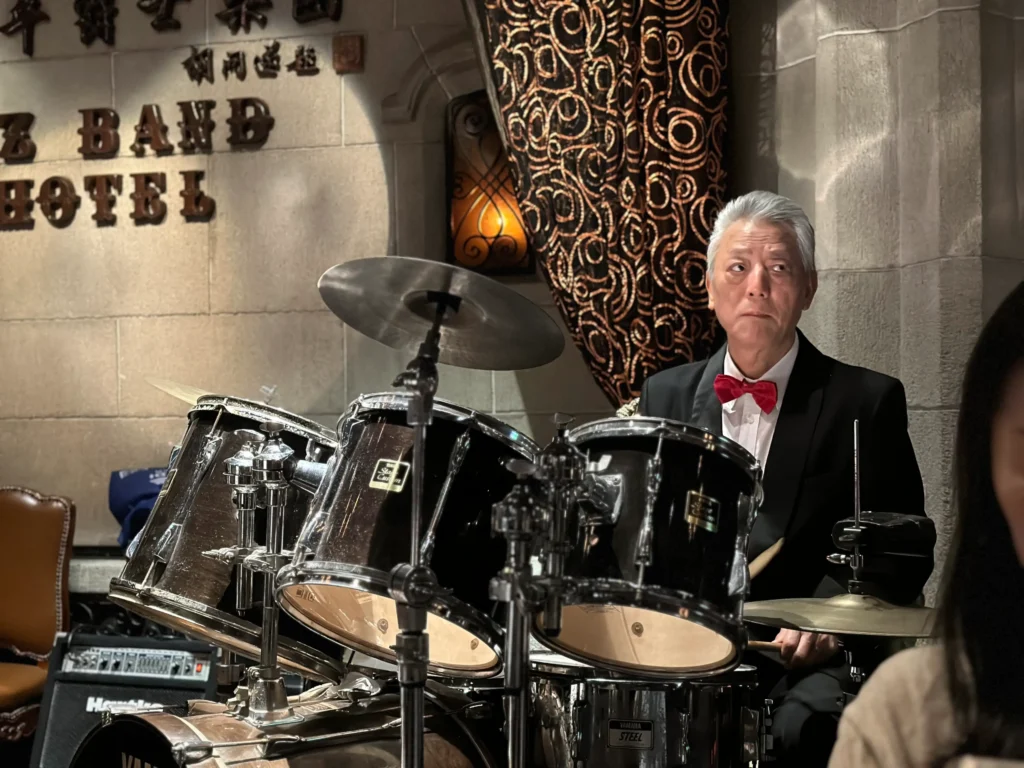 An older man in a tuxedo with a red bow tie sits behind a drum kit in a dimly lit, jazz-style venue. He gazes off to the side with a focused expression, positioned in front of a stone wall featuring elegant signage and patterned curtains.