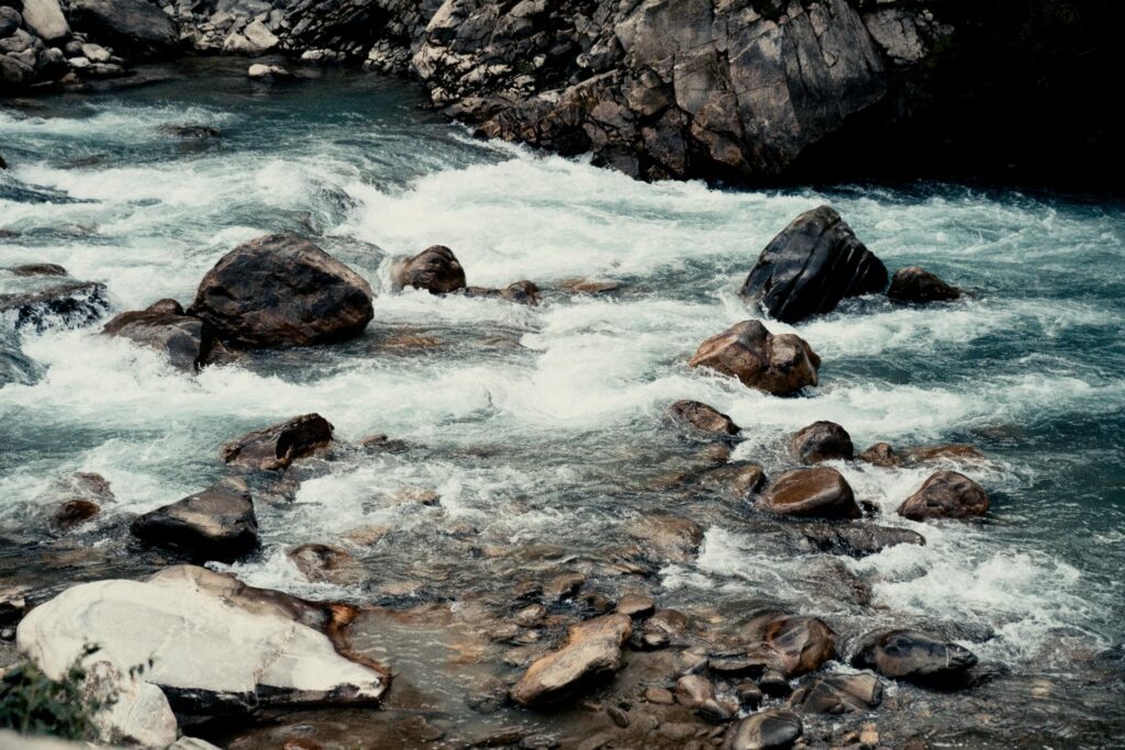 A turbulent river rushes over and around large, dark boulders nestled within a rocky canyon. The churning white water creates a sense of rapid movement against the stillness of the surrounding stone.