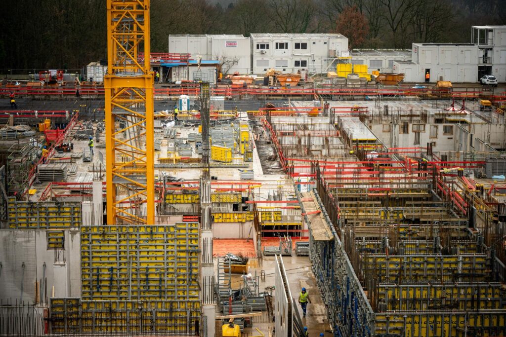 This high-angle shot captures a busy construction site filled with yellow crane segments, metal scaffolding, and concrete formwork. Workers in safety vests navigate the complex grid of building materials under an overcast sky.