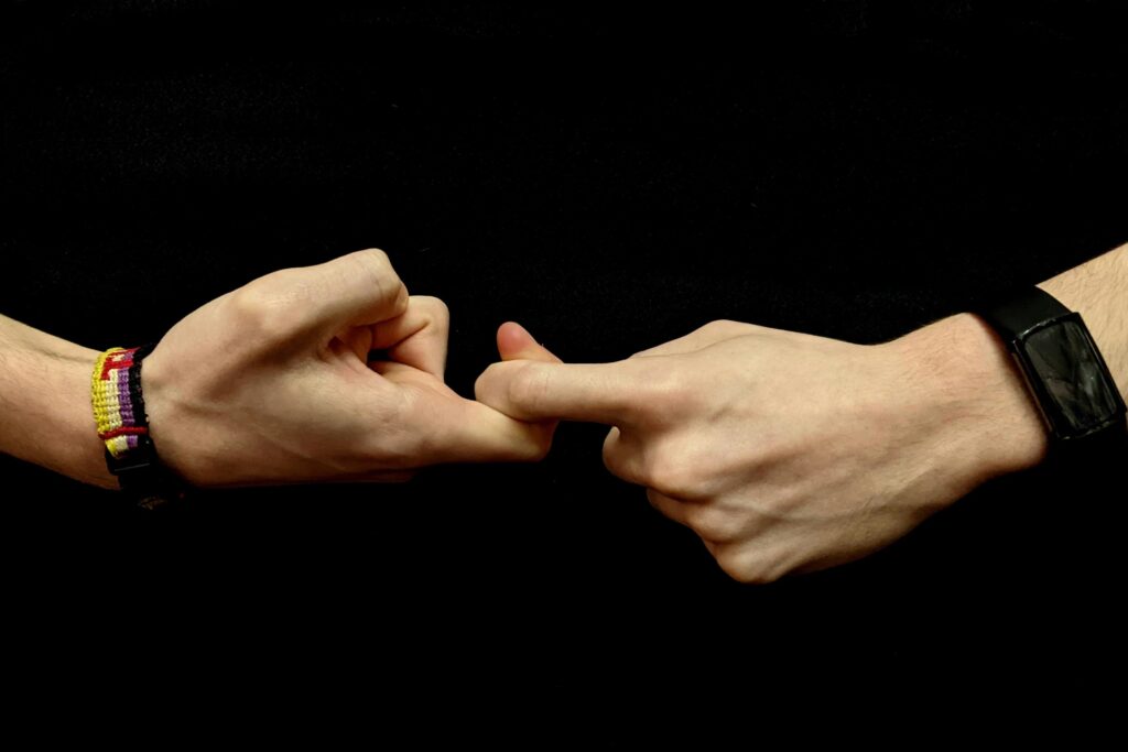 Two hands are shown making an interlocking gesture against a solid black background. One wrist wears a colorful woven bracelet while the other features a black digital watch.