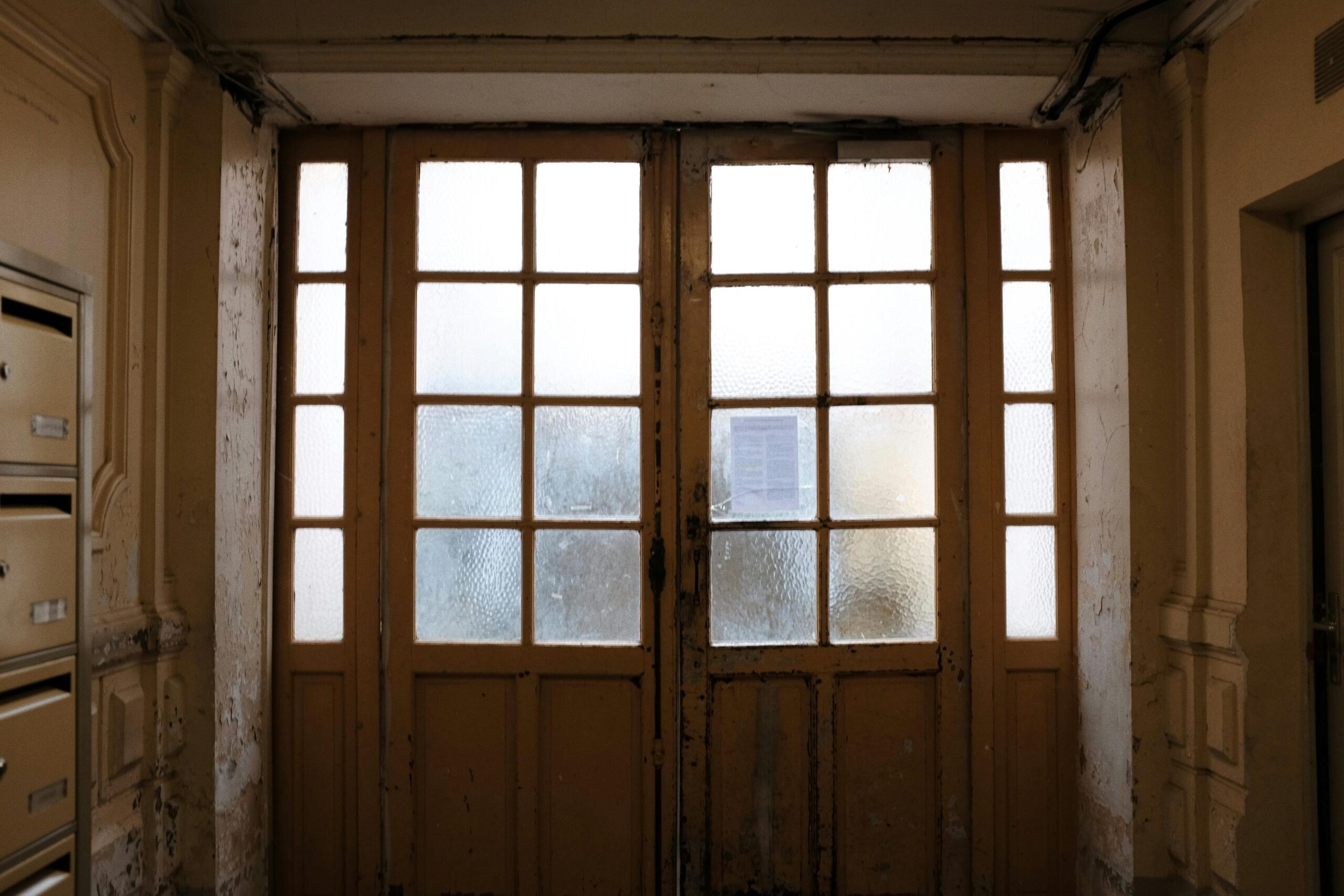 A pair of large, weathered wooden doors with glass panes stands at the end of a dim hallway. To the left, a stack of metal mailboxes is built into the aging, textured wall.