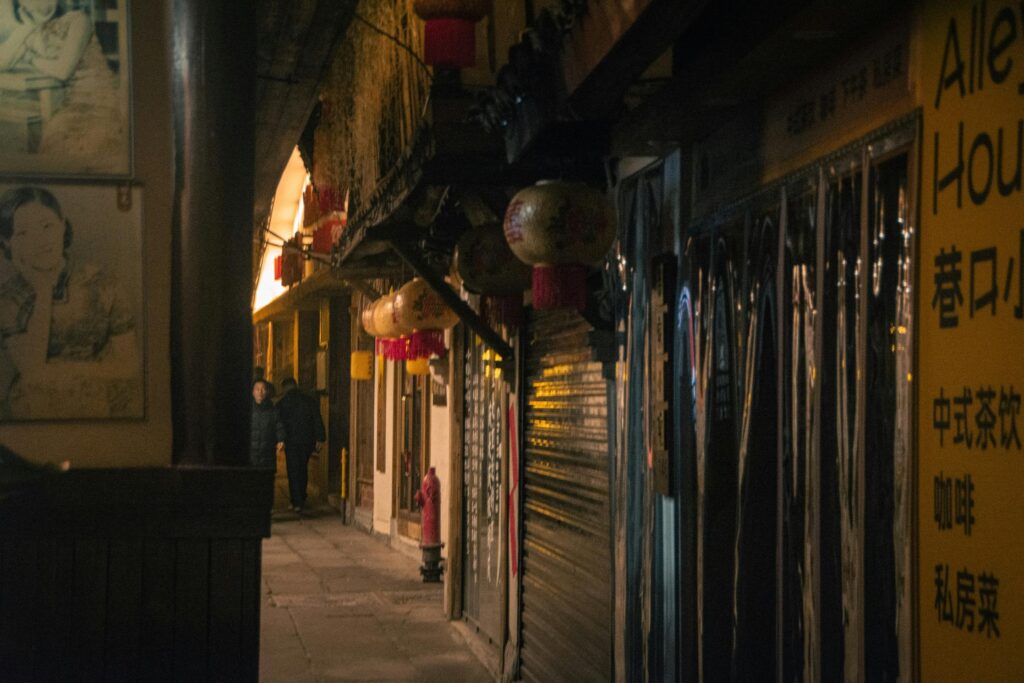 Warm light from traditional lanterns glows within a narrow, atmospheric alleyway lined with closed storefronts and vintage posters. The scene captures a quiet moment in a city at night, featuring a grainy texture and two figures walking into the distance.