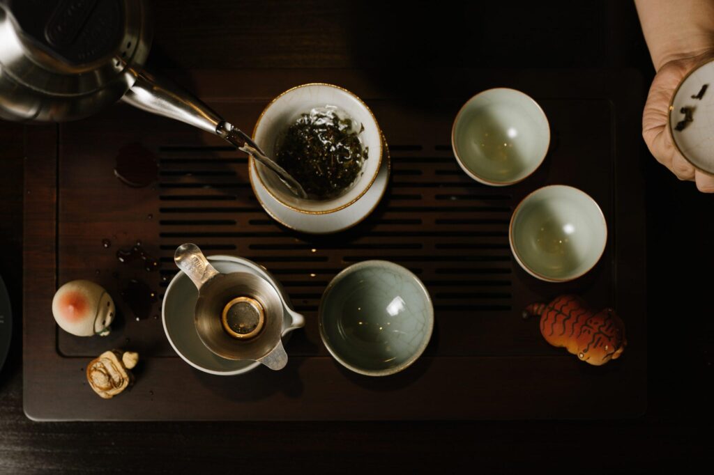 A person pours hot water from a kettle into a gaiwan filled with loose tea leaves. The tea set is arranged on a wooden slatted tray alongside small cups and decorative tea pets.