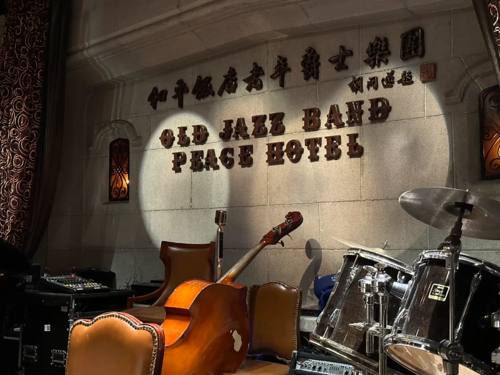 Musical instruments, including a drum set and a double bass, sit under spotlights in an elegant, stone-walled venue. The background features raised lettering for the "Old Jazz Band" at the "Peace Hotel."