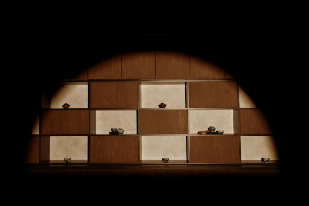 A wooden shelving unit with a checkerboard pattern of lit and dark compartments displays several small ceramic vessels. The entire scene is bathed in a warm, circular spotlight that fades into deep black surroundings.