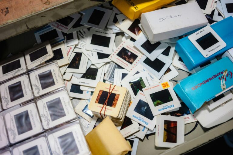 This image shows a cluttered overhead view of various vintage 35mm film slides, some loose and others organized in boxes or rubber-banded stacks. The collection includes notable brands like Kodak Ektachrome and Kodachrome, scattered alongside small storage containers in a nostalgic, disorganized pile.