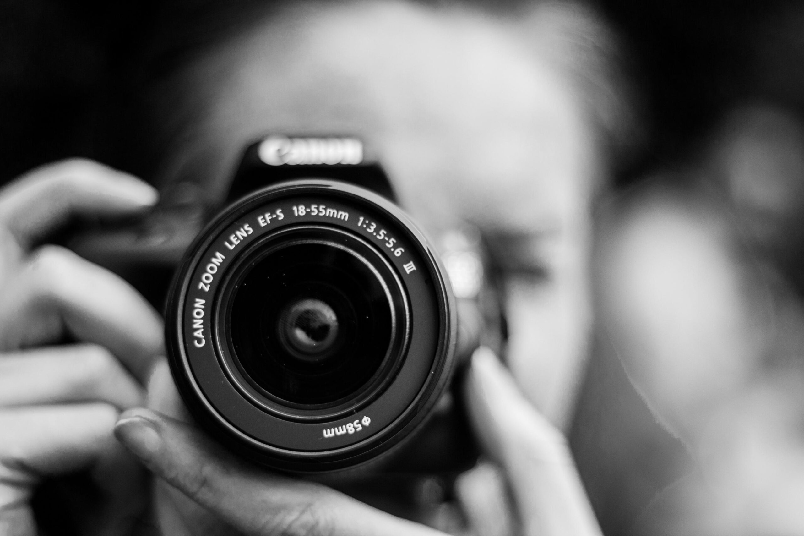A person holds a Canon DSLR camera directly toward the viewer, with the lens serving as the sharp focal point of the composition. The black-and-white photograph uses a shallow depth of field to blur the photographer’s face and background into a soft, textured bokeh.