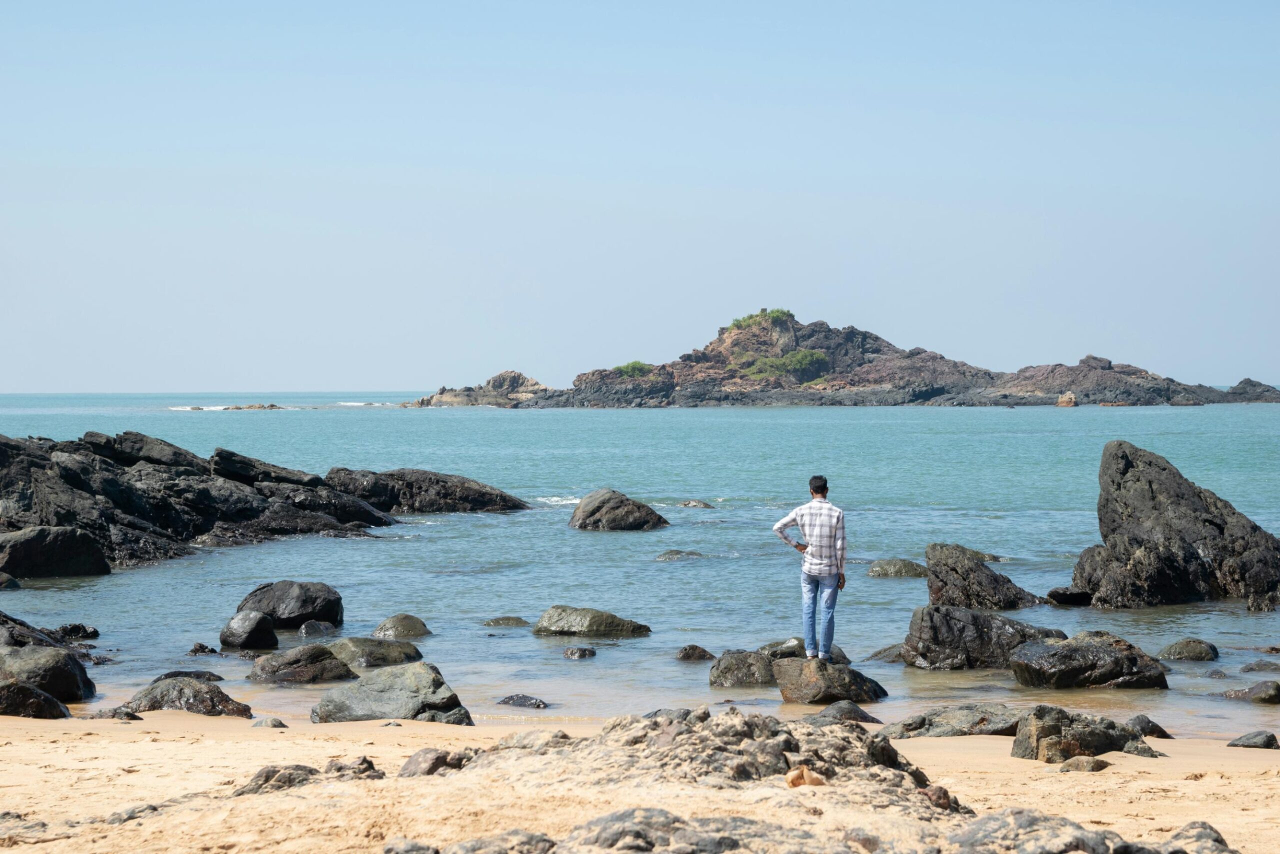 A person stands on dark rocks at the edge of a turquoise sea, looking out toward a rugged island in the distance. The scene is set against a clear blue sky on a bright, sandy beach.