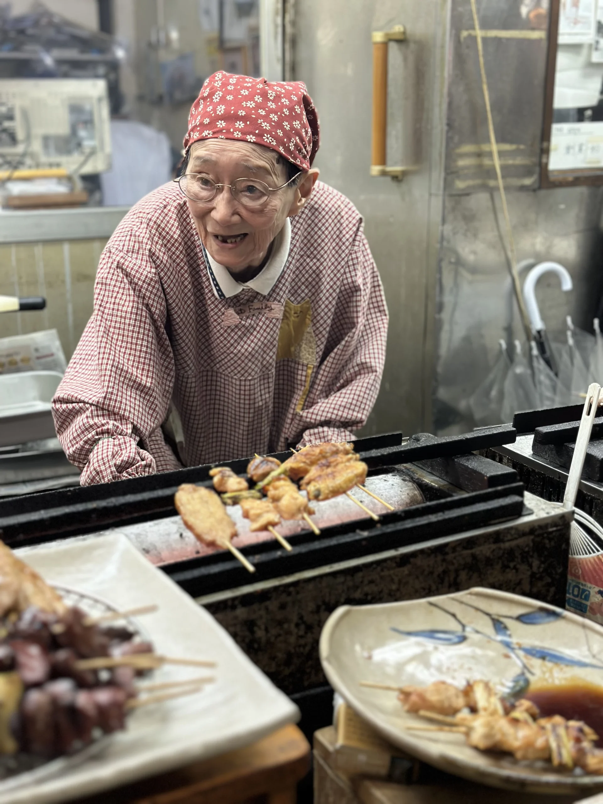 An elderly Japanese woman wearing a red floral headscarf and checkered apron grilling yakitori skewers over a charcoal fire at a street food stall.