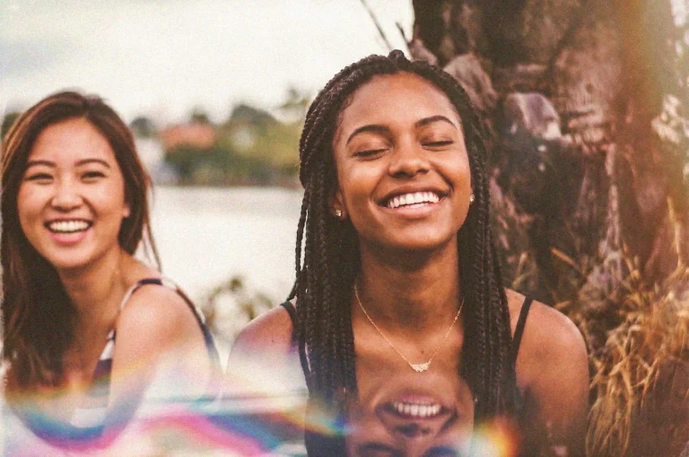 Two smiling young women outdoors with a vintage film effect, featuring a Black woman with braids and an East Asian woman.