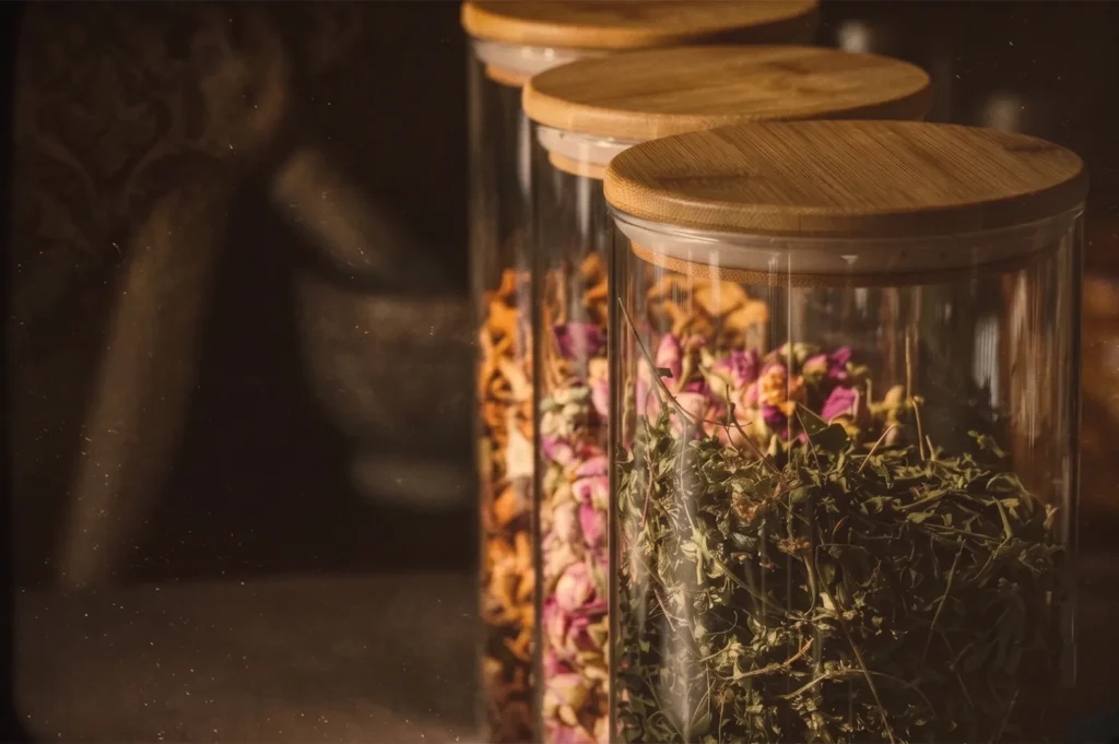 Three glass spice jars with bamboo lids containing dried herbs and rosebuds in a warm, dimly lit kitchen.