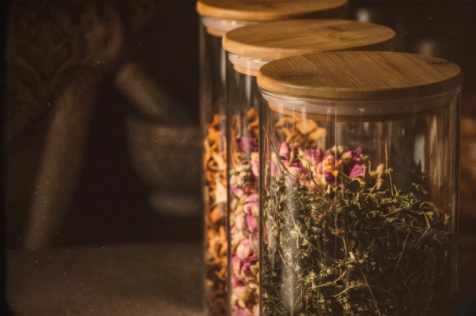 Three glass spice jars with bamboo lids containing dried herbs and rosebuds in a warm, dimly lit kitchen.