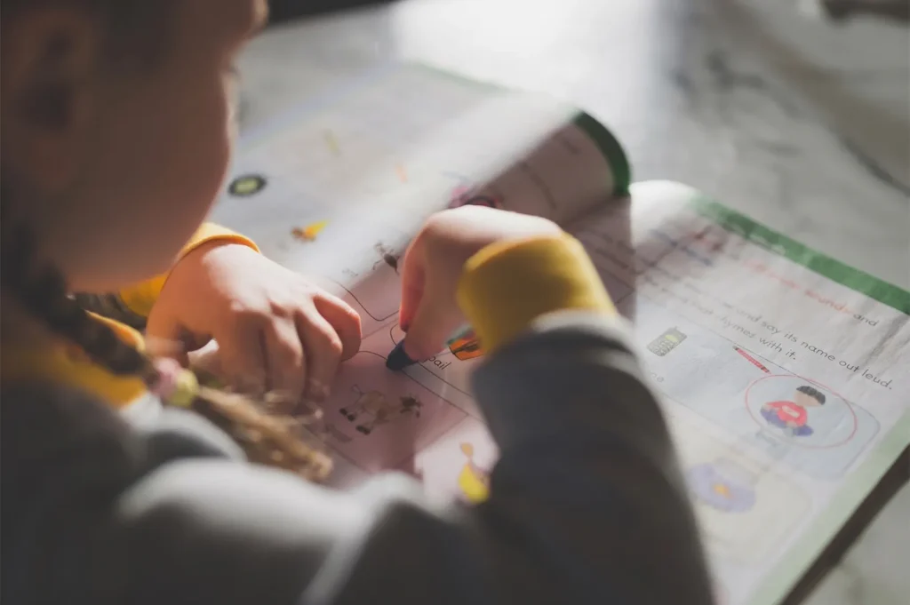 Young girl with braided hair using a crayon to complete an educational workbook activity.