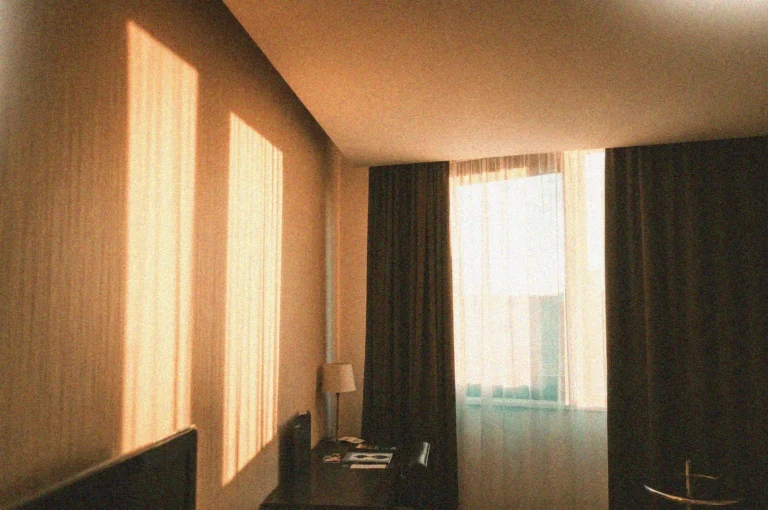 Golden hour sunlight streaming through sheer curtains into a dark, modern hotel room or bedroom.