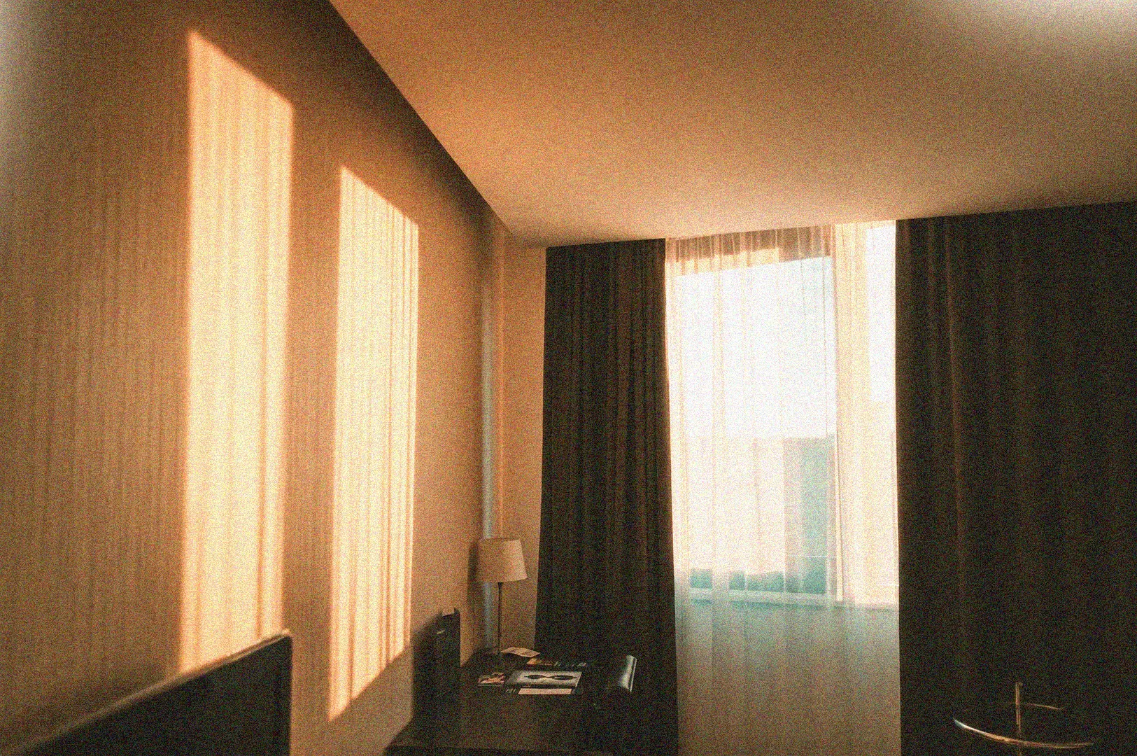 Golden hour sunlight streaming through sheer curtains into a dark, modern hotel room or bedroom.