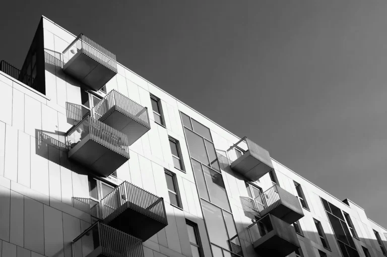 Black and white low-angle shot of a modern apartment building featuring geometric balconies and sharp shadows against a clear sky.