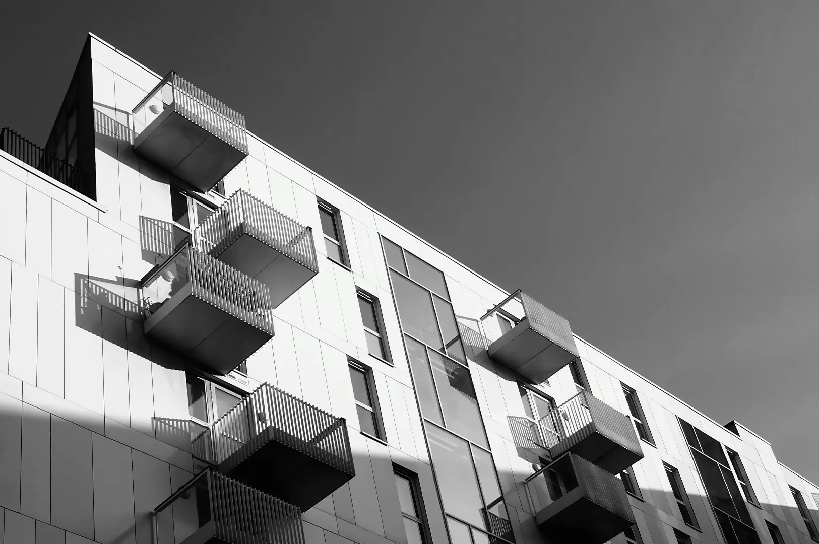Black and white low-angle shot of a modern apartment building featuring geometric balconies and sharp shadows against a clear sky.