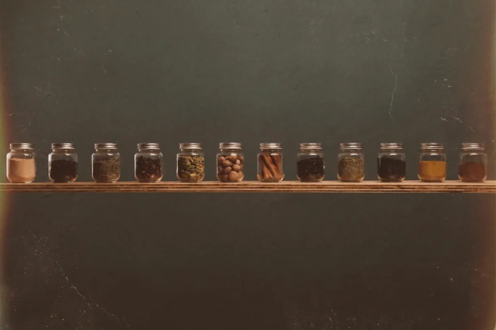 A row of small glass jars filled with various colorful spices and herbs on a wooden shelf against a dark, textured wall.