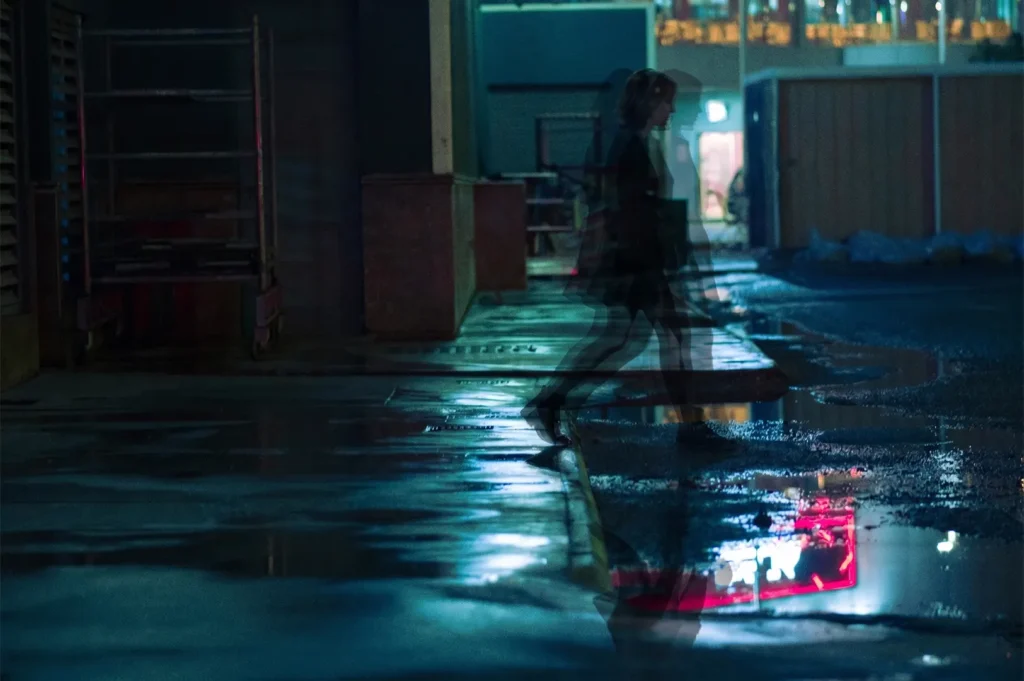 Double exposure of a woman walking down a dark city alley at night with wet ground reflections.