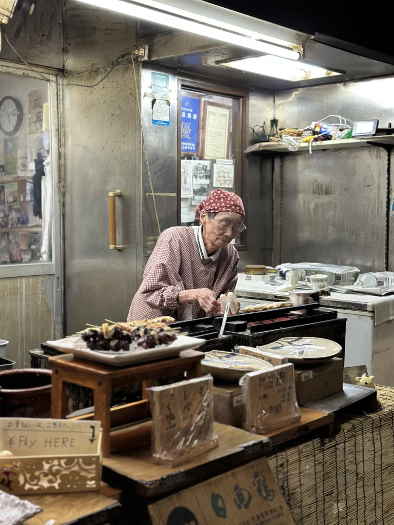 Elderly Japanese street food vendor in a red headscarf preparing grilled yakitori skewers at a traditional stall with a "Pay Here" sign.