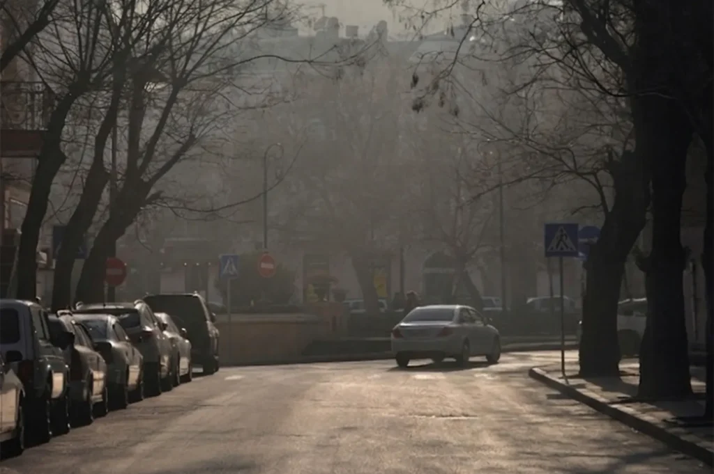 A white car driving down a hazy city street lined with parked cars and bare winter trees.