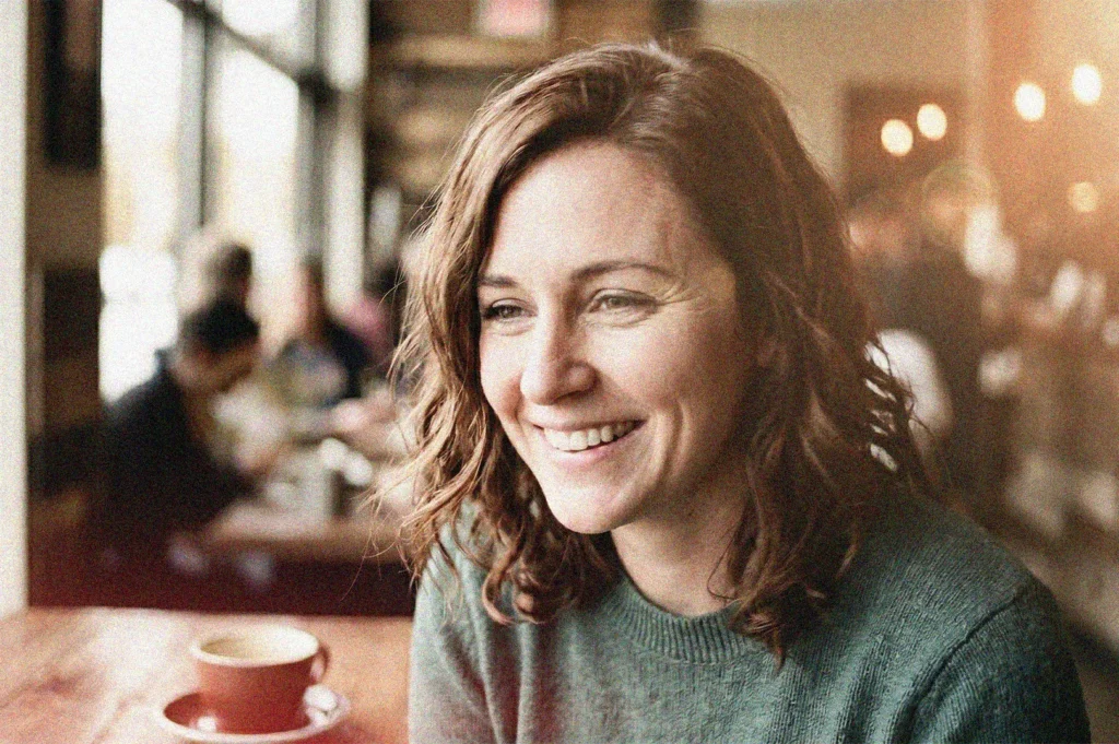 A smiling woman with wavy brown hair sitting at a table in a sunlit cafe with a coffee cup in the foreground.