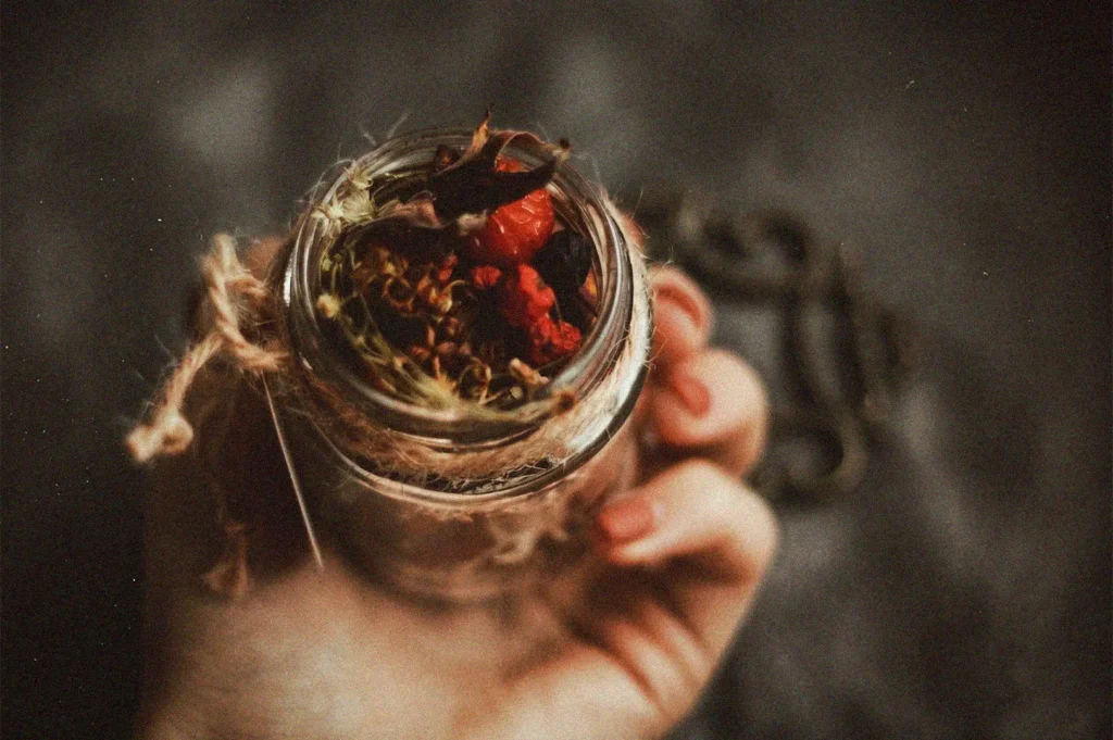 A hand holding a small glass jar filled with colorful dried spices and rosebuds, decorated with a rustic twine bow.