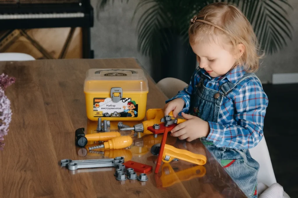 A young girl in denim overalls playing with a yellow plastic toy tool set at a wooden table.