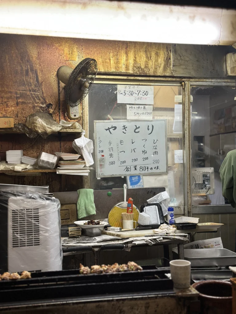 Interior of a rustic Japanese yakitori stall featuring a handwritten menu board, a small charcoal grill with chicken skewers, and a vintage electric fan.