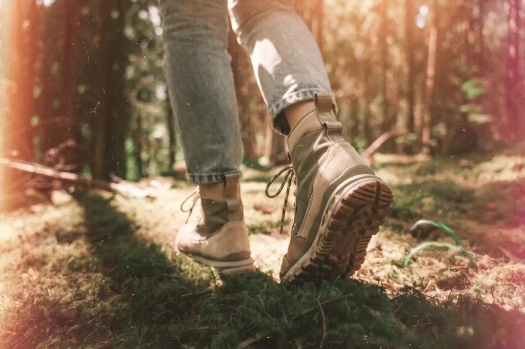 A person wearing denim jeans and beige hiking boots walks along a moss-covered path through a sunlit forest.