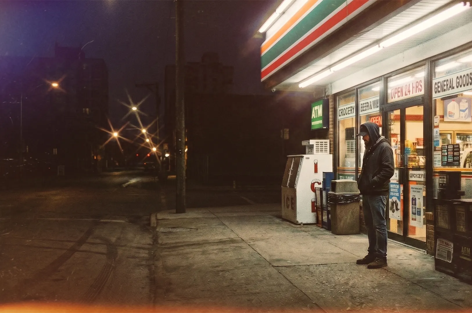 A man in a hooded jacket standing outside a 7-Eleven convenience store at night on an empty city street.