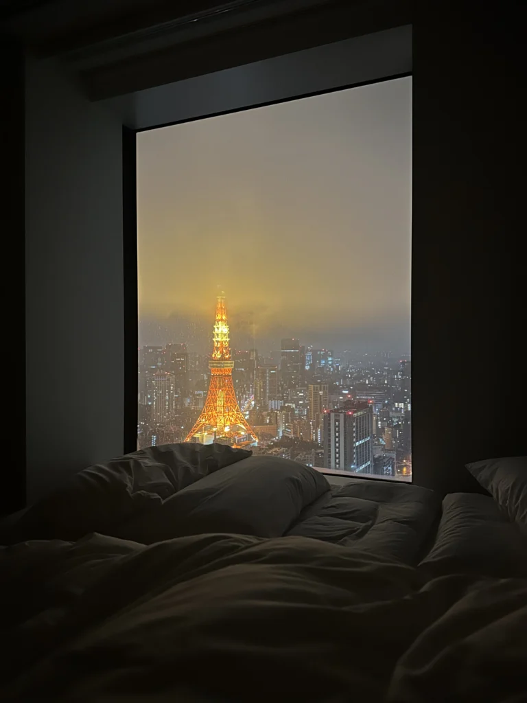 An atmospheric night view of the illuminated Tokyo Tower framed by a hotel window, overlooking a foggy city skyline from a dark bedroom with unmade white linens.