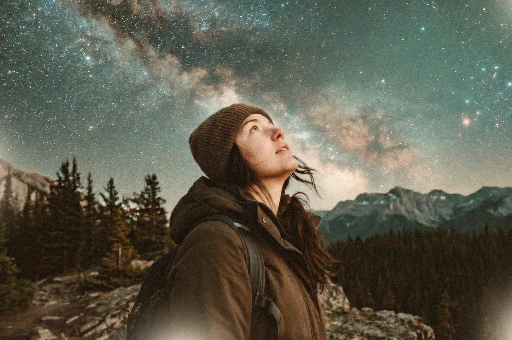 Close-up of a female hiker wearing a winter beanie and jacket, looking up at the starry Milky Way night sky over a mountain forest.