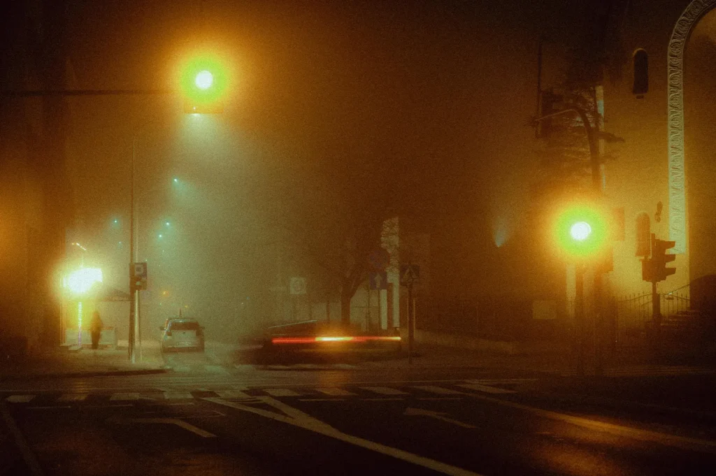 A long-exposure shot of a foggy city street at night, with glowing yellow streetlights and blurred light trails from a passing car.