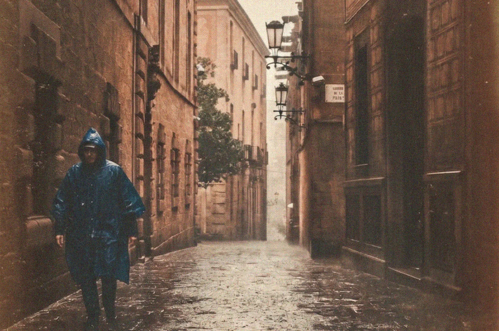 A person wearing a blue hooded raincoat walks down a wet, cobbled European street lined with historic stone buildings on a rainy day.