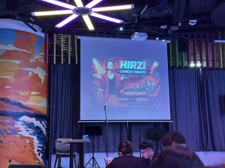 Hirzi Comedy Nights 2026 weekly stand-up show poster projected on a stage screen.