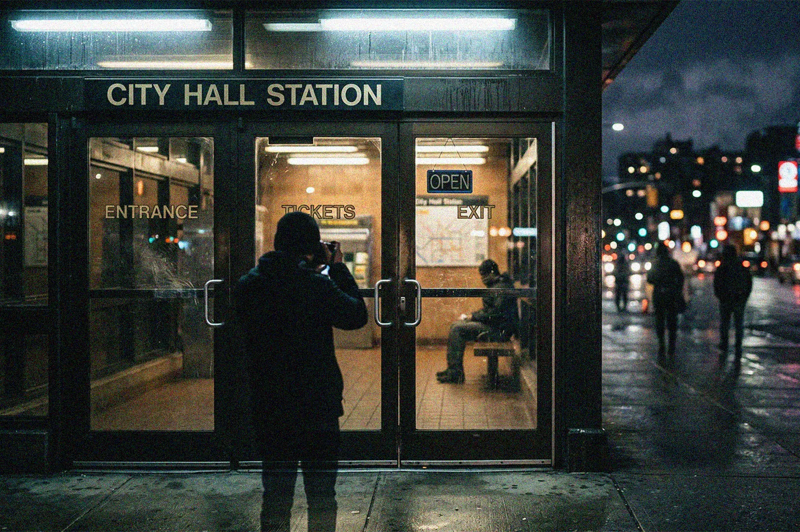 A photographer taking a picture outside the glass doors of a City Hall Station subway entrance on a rainy night.
