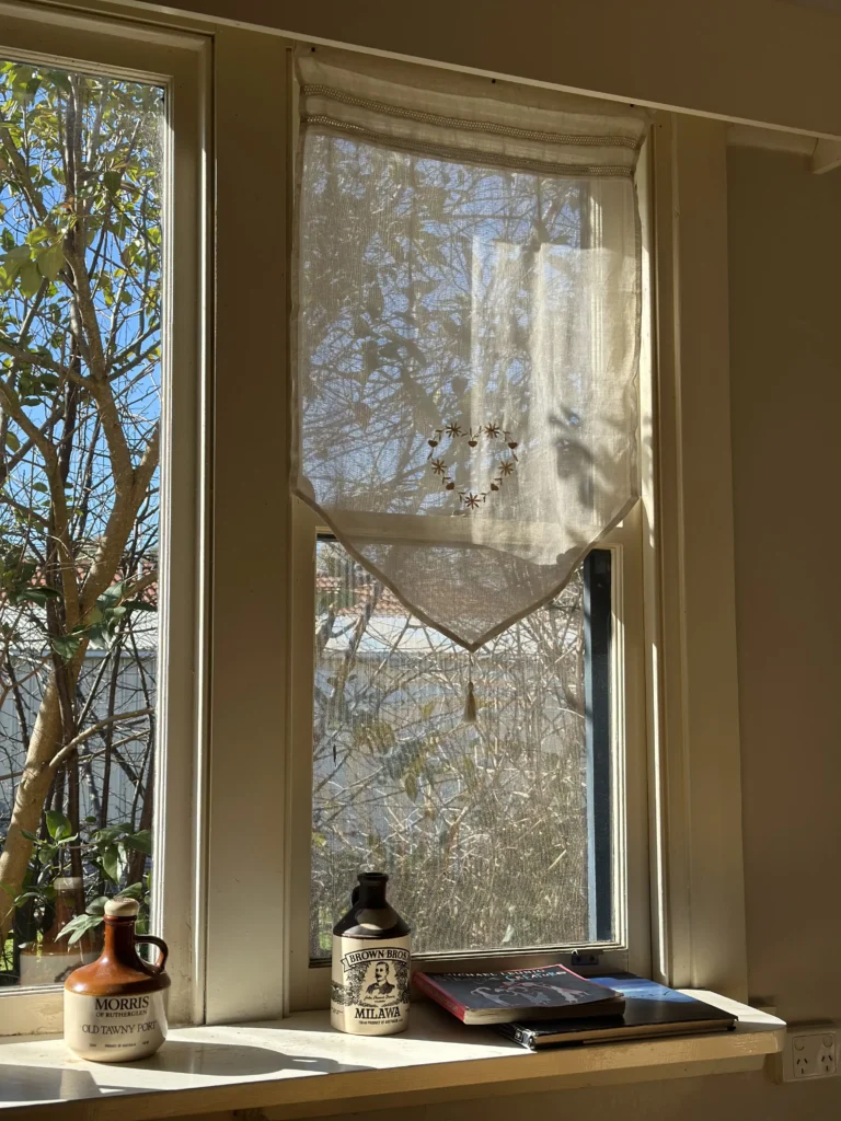 Vintage-style window sill with an embroidered sheer cafe curtain, antique brown stoneware bottles, and books in warm sunlight.