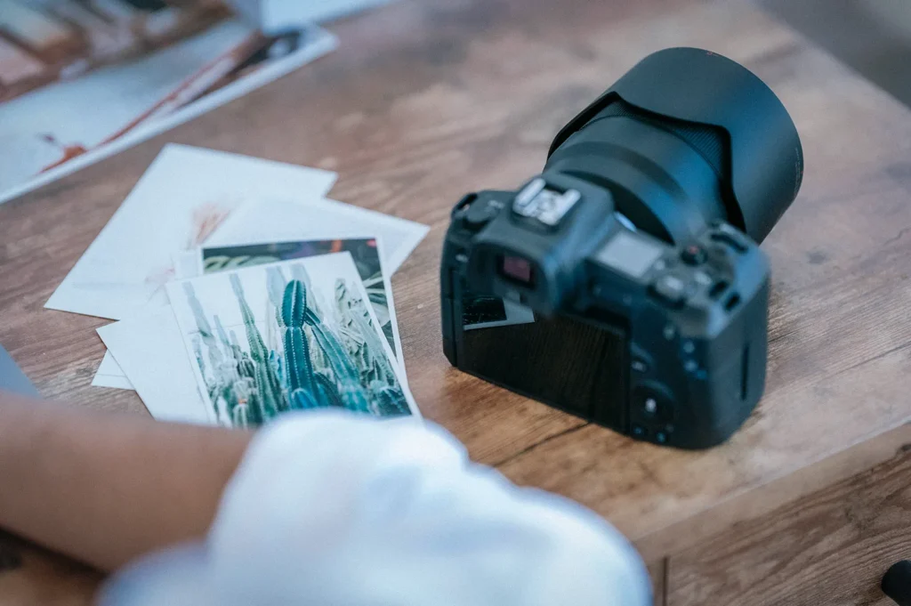 Mirrorless digital camera on a wooden table beside printed photographs, with a photographer reviewing image prints during a photo editing and photography workflow.