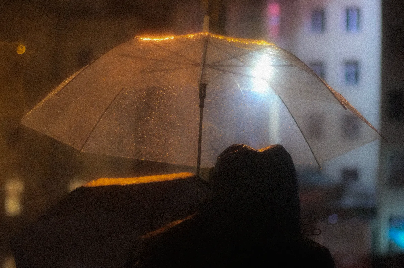 Person holding a transparent umbrella on a rainy night, illuminated by warm city lights with raindrops visible on the umbrella and blurred urban buildings in the background.