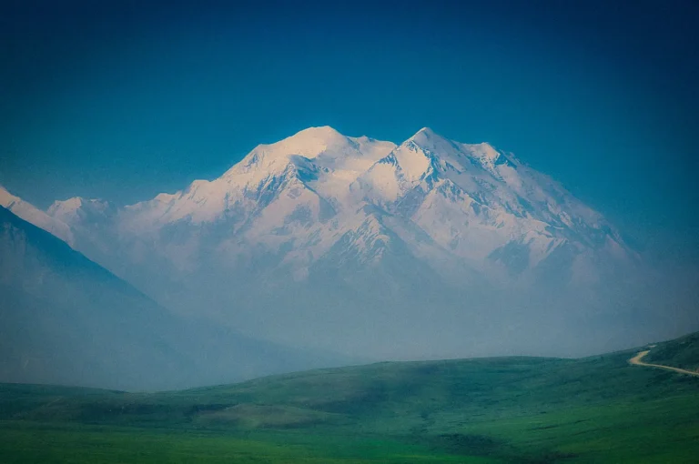 Snow-capped mountain range towering above a vast green valley under a clear blue sky, showcasing a scenic alpine landscape with rolling hills and distant peaks.