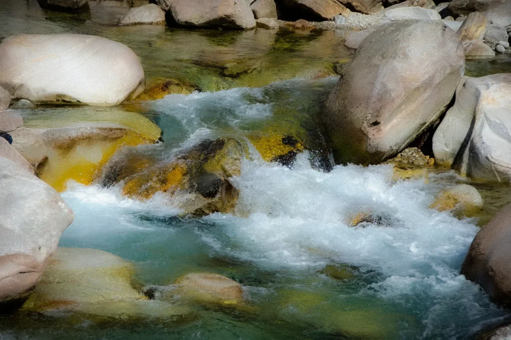 Crystal-clear mountain spring water flowing through smooth rocks and boulders, creating a fresh natural stream in a pristine outdoor landscape.