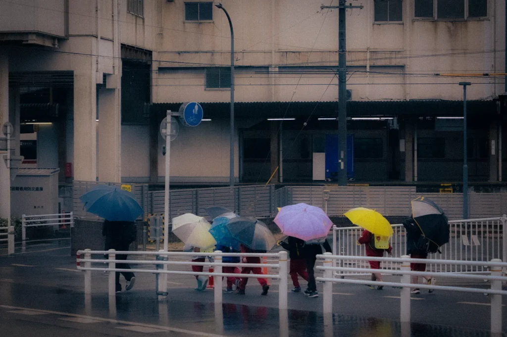 Pedestrians carrying colorful umbrellas while crossing a wet city street on a rainy day, with a train station platform and urban buildings in the background.