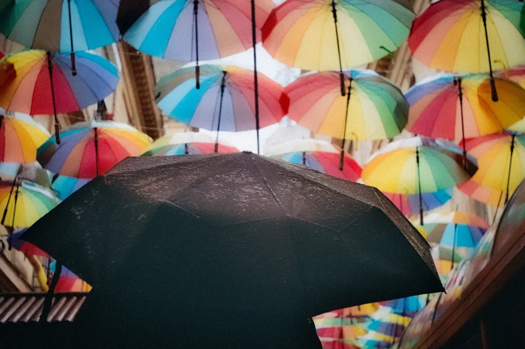 Colorful umbrella canopy installation suspended above a narrow street, featuring vibrant rainbow umbrellas and a person holding a black umbrella below during light rain.