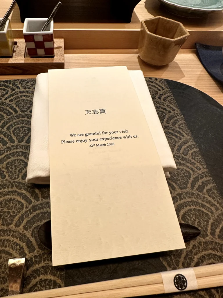 Elegant Japanese omakase place setting with a personalized welcome card, chopsticks, ceramic cup, and patterned placemat at a premium sushi counter.