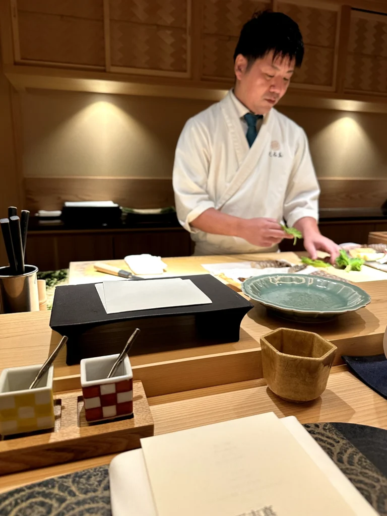 Japanese chef preparing ingredients behind a wooden omakase counter with ceramic tableware in an intimate fine dining restaurant.