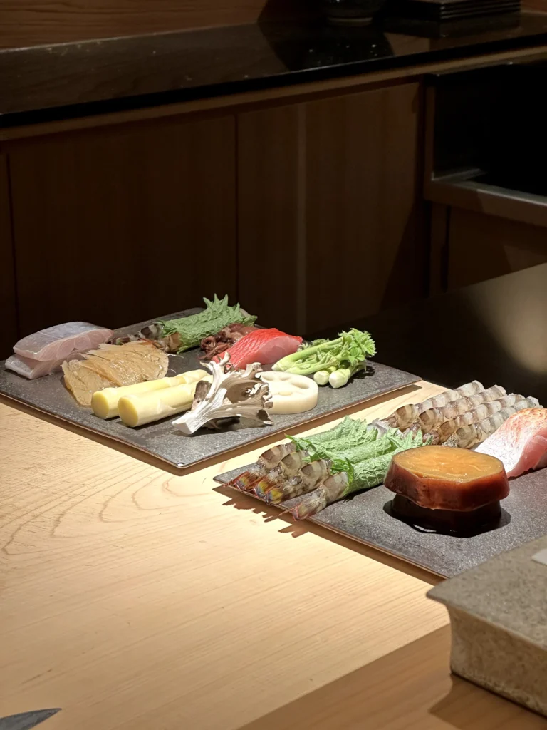 Premium omakase ingredients arranged on stone plates, featuring fresh seafood, prawns, lotus root, bamboo shoots, mushrooms, and seasonal vegetables at a Japanese sushi counter.