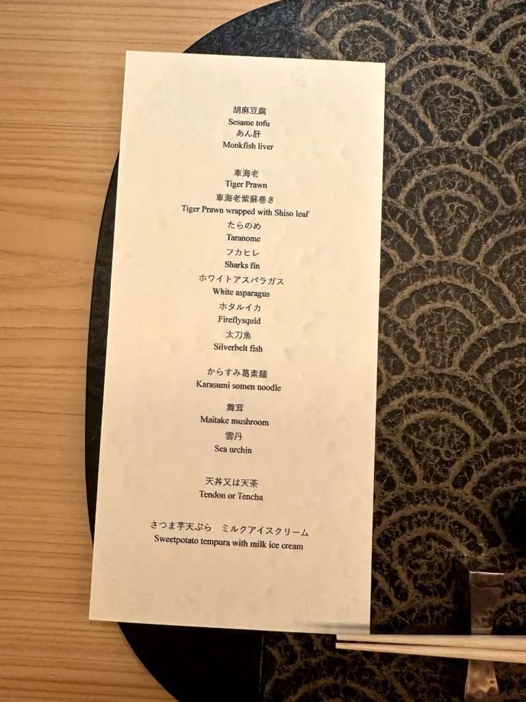 Omakase menu card listing seasonal Japanese dishes including sesame tofu, tiger prawn, shark fin, white asparagus, sea urchin, tendon or teaccha, and sweet potato tempura with milk ice cream.